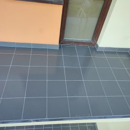 A Line interiors - Balkon z szarą, błyszczącą terakotą ułożoną w regularną siatkę, widok z góry na narożnik balkonu z oknem i pomarańczową ścianą.