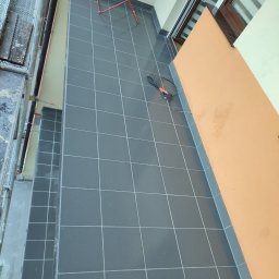 A Line interiors - Wykafelkowana podłoga balkonu w odcieniach szarości z widocznym pomarańczowym fragmentem ściany i rozłożoną suszarką na pranie, fragment rusztowania po lewej stronie.