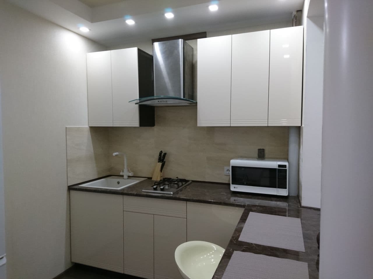 Jasna kuchnia z białymi szafkami górnymi i dolnymi, szarym blatem, płytkami imitującymi kamień na ścianie, srebrnym okapem i białą mikrofalówką, oświetlona światłem punktowym.