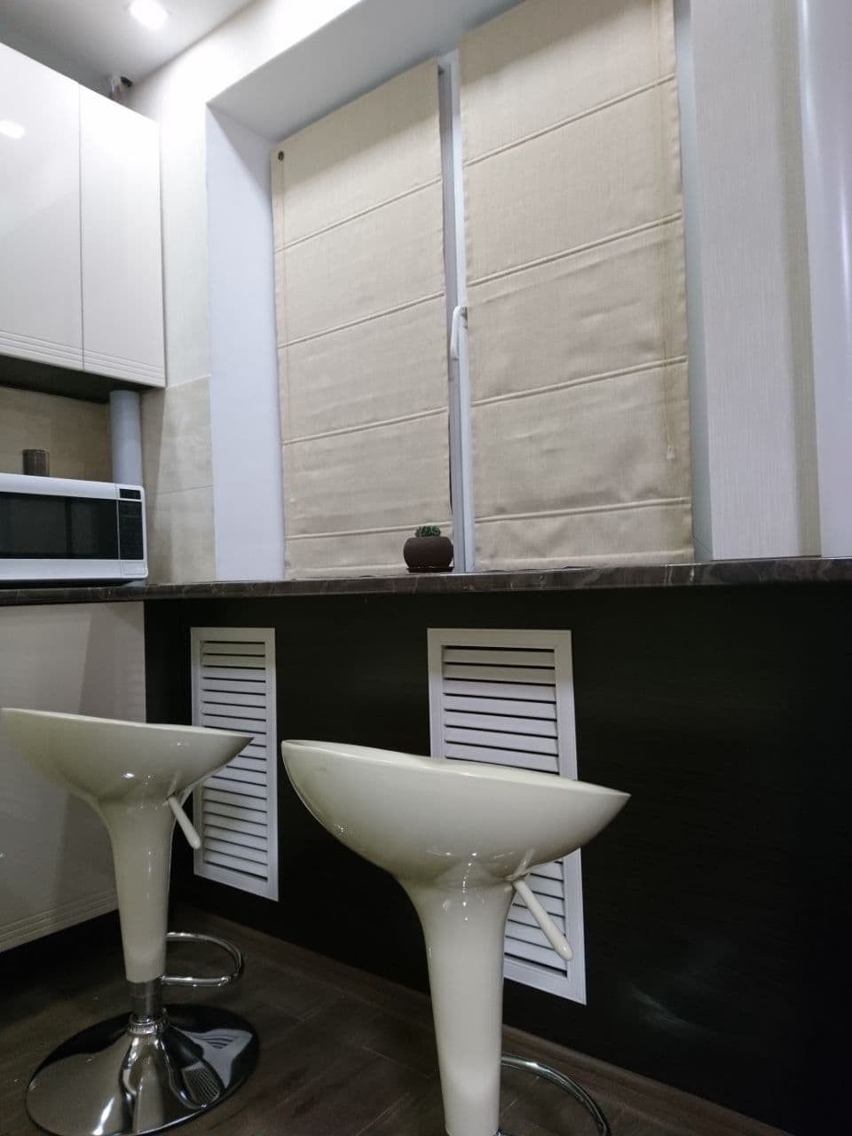 Nowoczesna kuchnia z białymi szafkami, dwoma białymi hokerami i beżowymi roletami rzymskimi w oknie, widok z niskiego kąta.