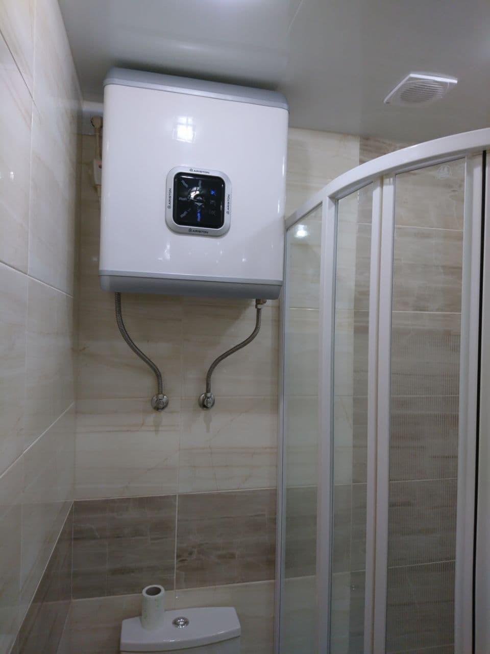 Kompaktowy, biały bojler Ariston zamontowany nad toaletą w łazience wykończonej w beżowych i brązowych płytkach, obok kabiny prysznicowej z przesuwnymi drzwiami.