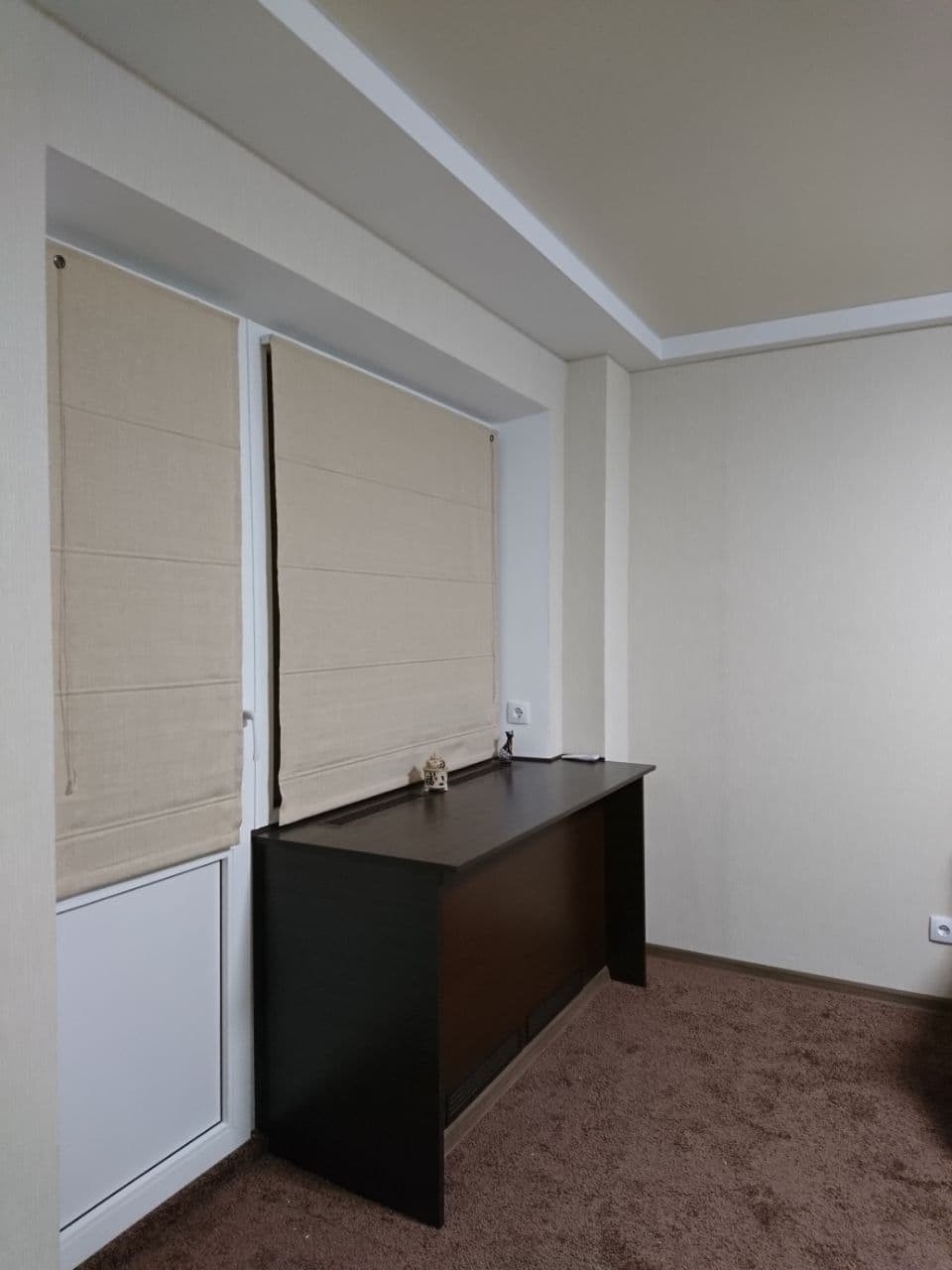 Beżowe rolety rzymskie zamontowane w oknie balkonowym, z ciemnobrązowym biurkiem przylegającym do ramy okiennej, na tle beżowych ścian i brązowej wykładziny dywanowej.