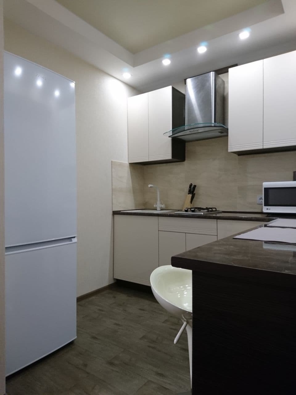 Jasna kuchnia z białymi szafkami, okapem ze stali nierdzewnej, białym krzesłem z tworzywa sztucznego i podłogą imitującą ciemne drewno.