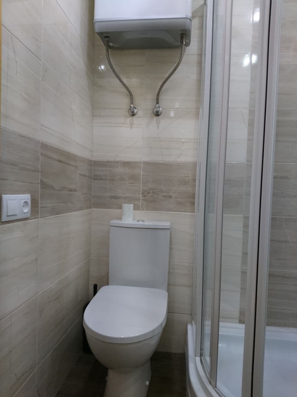 Ukończona łazienka z toaletą, kabiną prysznicową i podgrzewaczem wody na ścianie wyłożonej płytkami w odcieniach beżu i brązu.