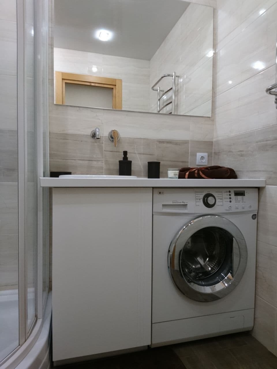 Nowoczesna łazienka z pralką zabudowaną pod umywalką, jasne płytki imitujące drewno, duże lustro z odbiciem drzwi i grzejnika ręcznikowego.