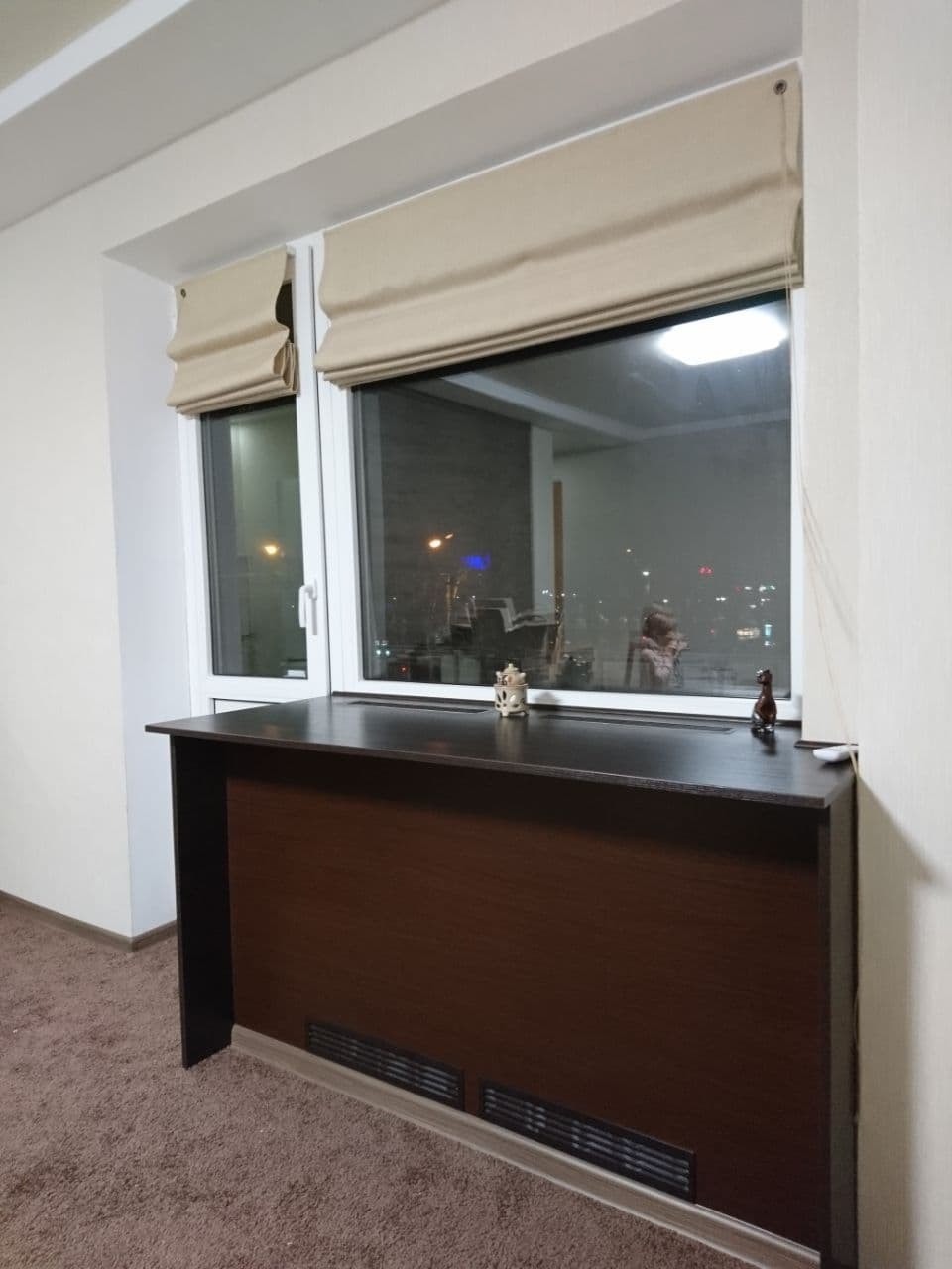 Balkon z zabudową okienną i brązowym blatem, ozdobiony figurkami, z beżowymi roletami rzymskimi.