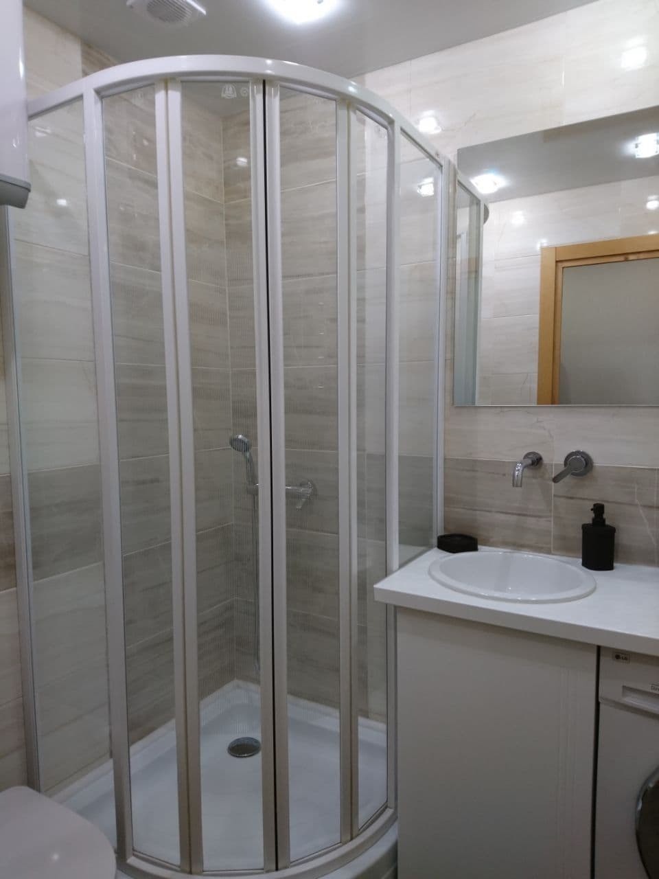 Narożna kabina prysznicowa z transparentnymi drzwiami w łazience wykończonej płytkami w odcieniach beżu, widoczna umywalka nablatowa z czarnym dozownikiem mydła i bateria umywalkowa.