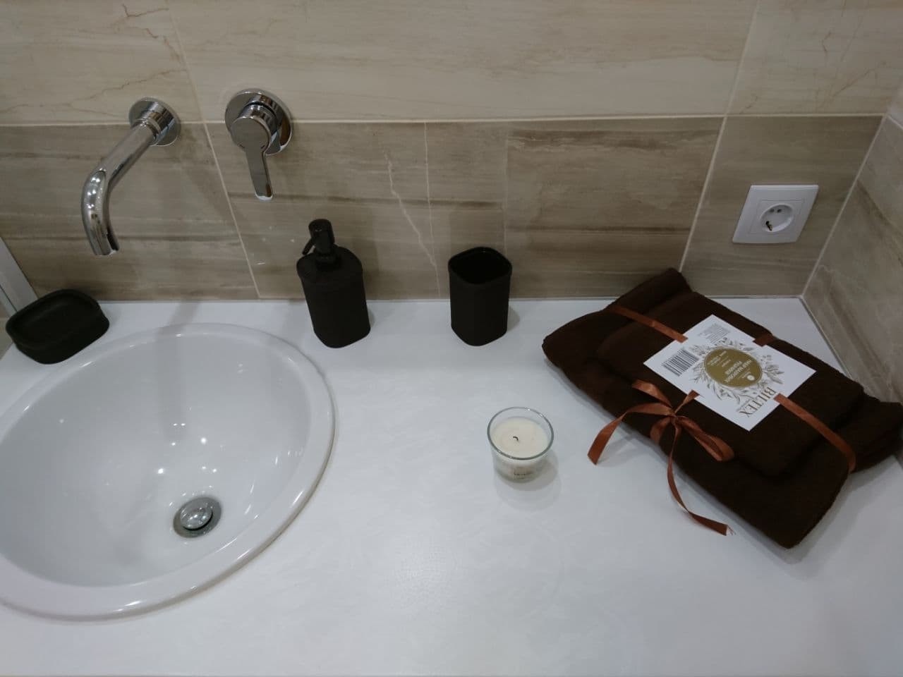 Umywalka w łazience z chromowaną baterią, czarnym dozownikiem mydła, kubkiem i zestawem brązowych ręczników z etykietą Biletex oraz zapaloną świeczką na blacie z beżowymi płytkami na ścianie.