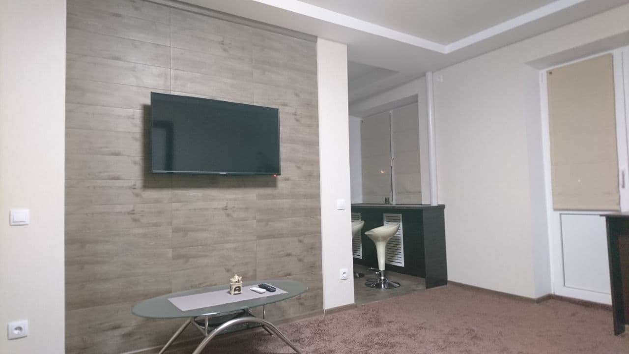 Telewizor zawieszony na ścianie z płytek imitujących drewno w salonie z aneksem kuchennym.