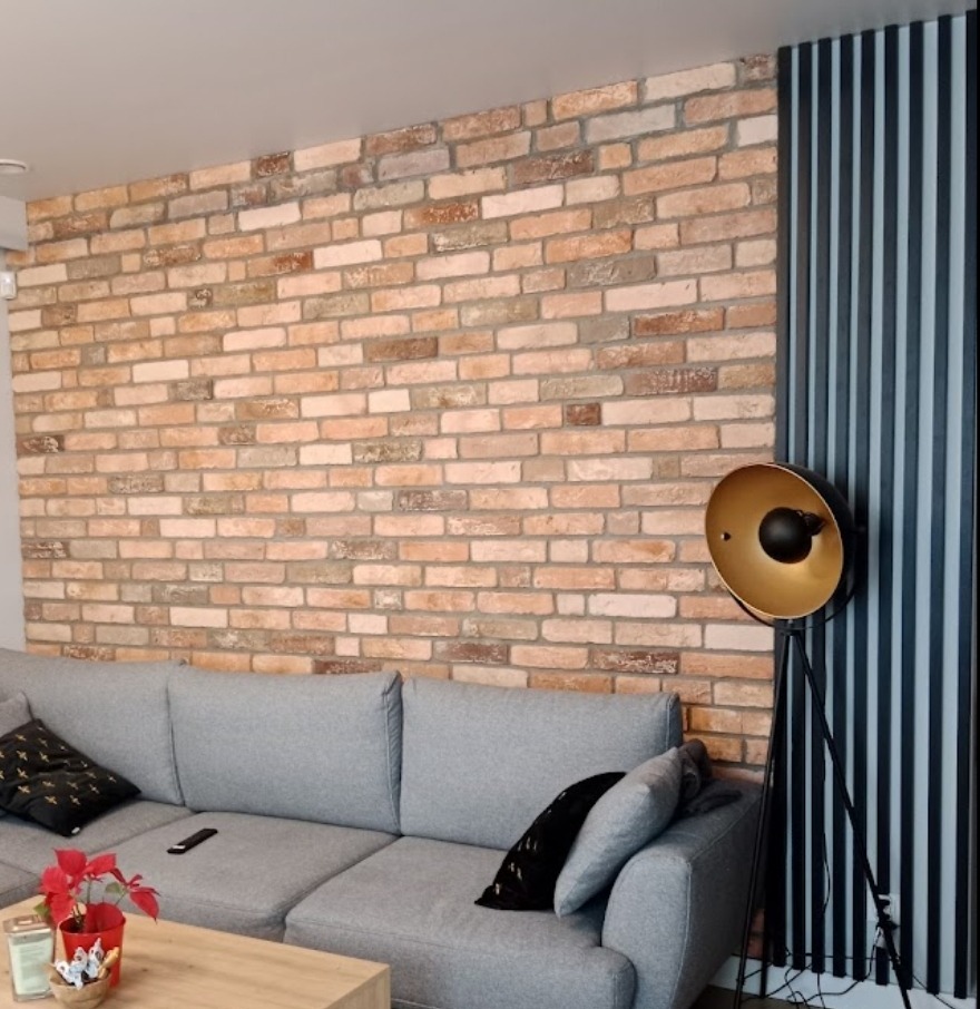 Ceglana ściana z dekoracyjnymi panelami pionowymi w kolorze grafitowym, szara sofa z poduszkami i lampa stojąca z kloszem w kolorze złota.