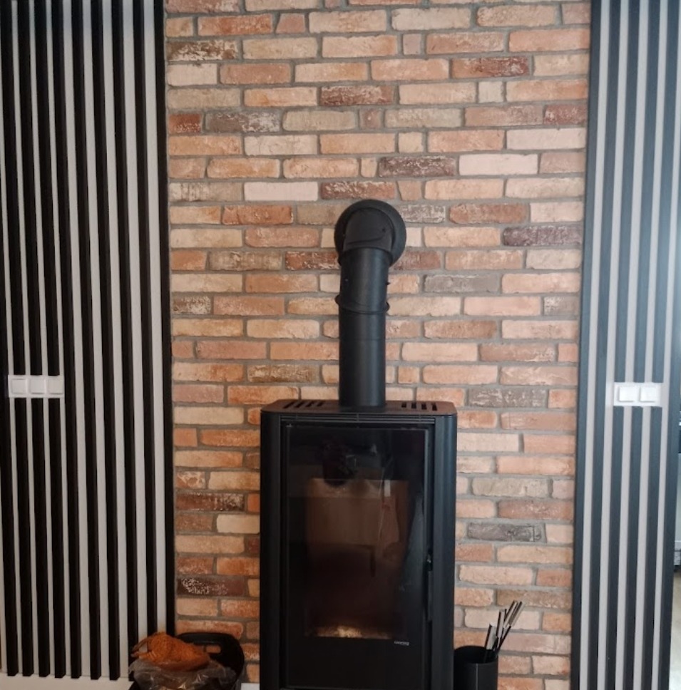 Ceglana ściana z kominkiem w nowoczesnym salonie, czarne pionowe panele dekoracyjne po bokach, widoczne akcesoria kominkowe.