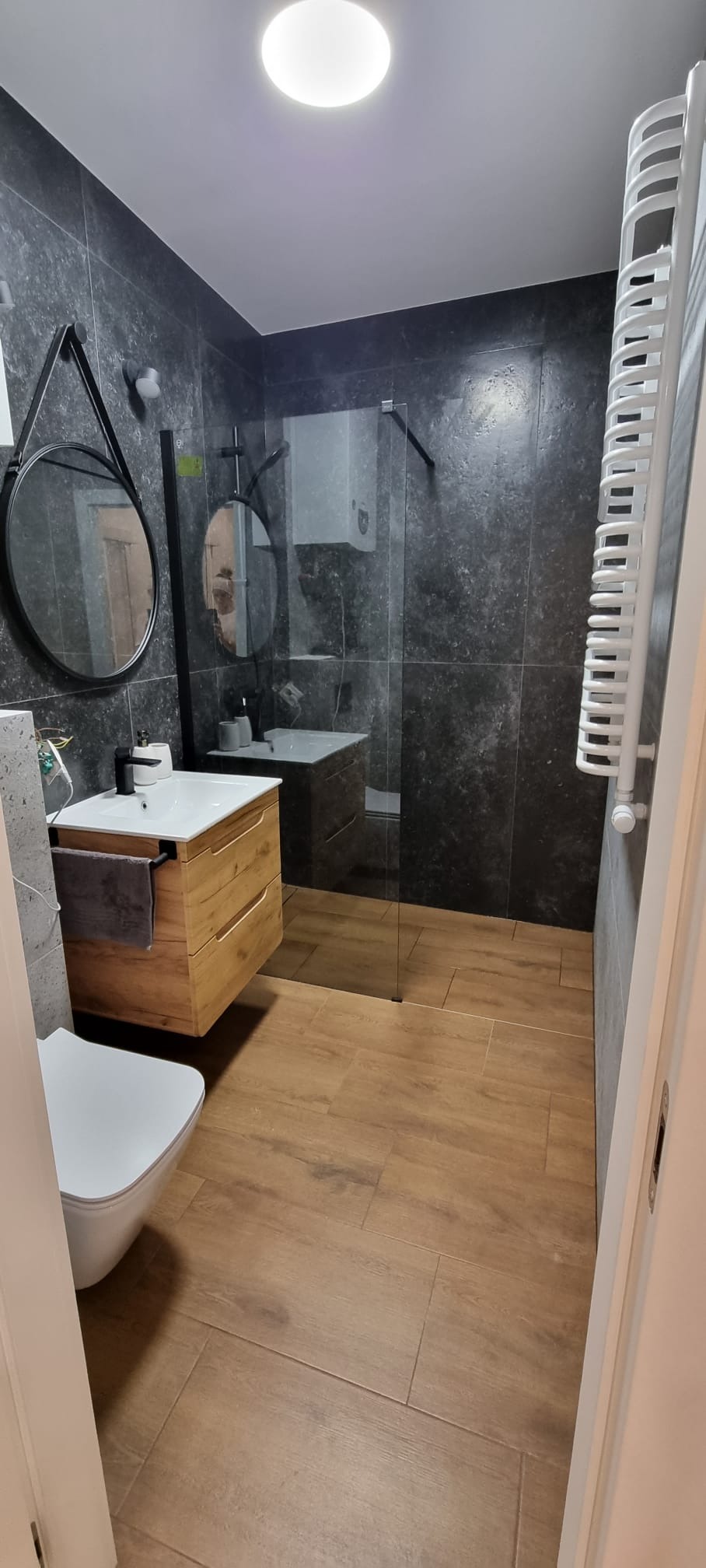 Nowoczesna łazienka z grafitowymi płytkami imitującymi kamień, drewnopodobną podłogą, wiszącą szafką z umywalką, okrągłym lustrem na pasku i kabiną prysznicową typu walk-in.