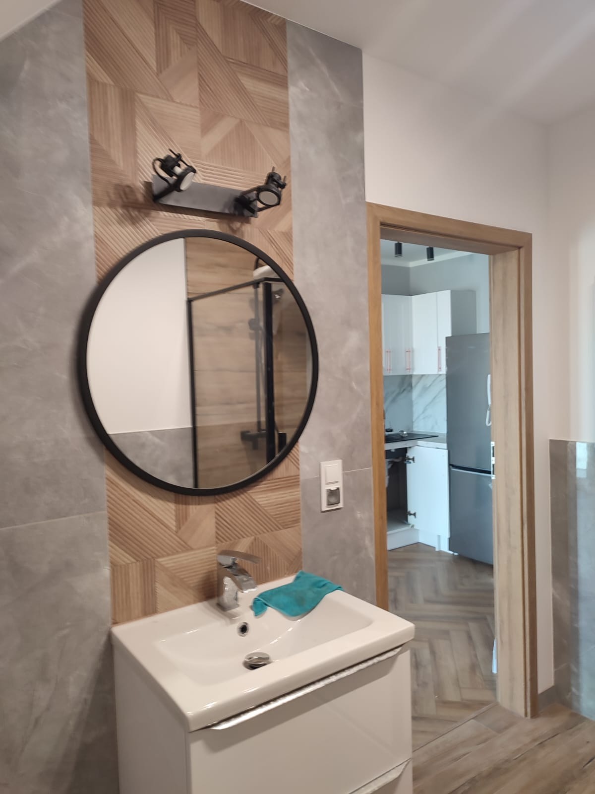 Nowoczesna łazienka z okrągłym lustrem w czarnej ramie, umywalką na szafce, designerską lampą nad lustrem i widokiem na kuchnię w tle.