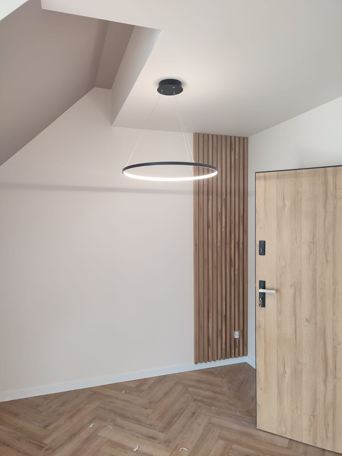 Wykończone wnętrze: parkiet w jodełkę, drewniana boazeria pionowa, drzwi wewnętrzne z klamką i okrągła lampa sufitowa z białym światłem.
