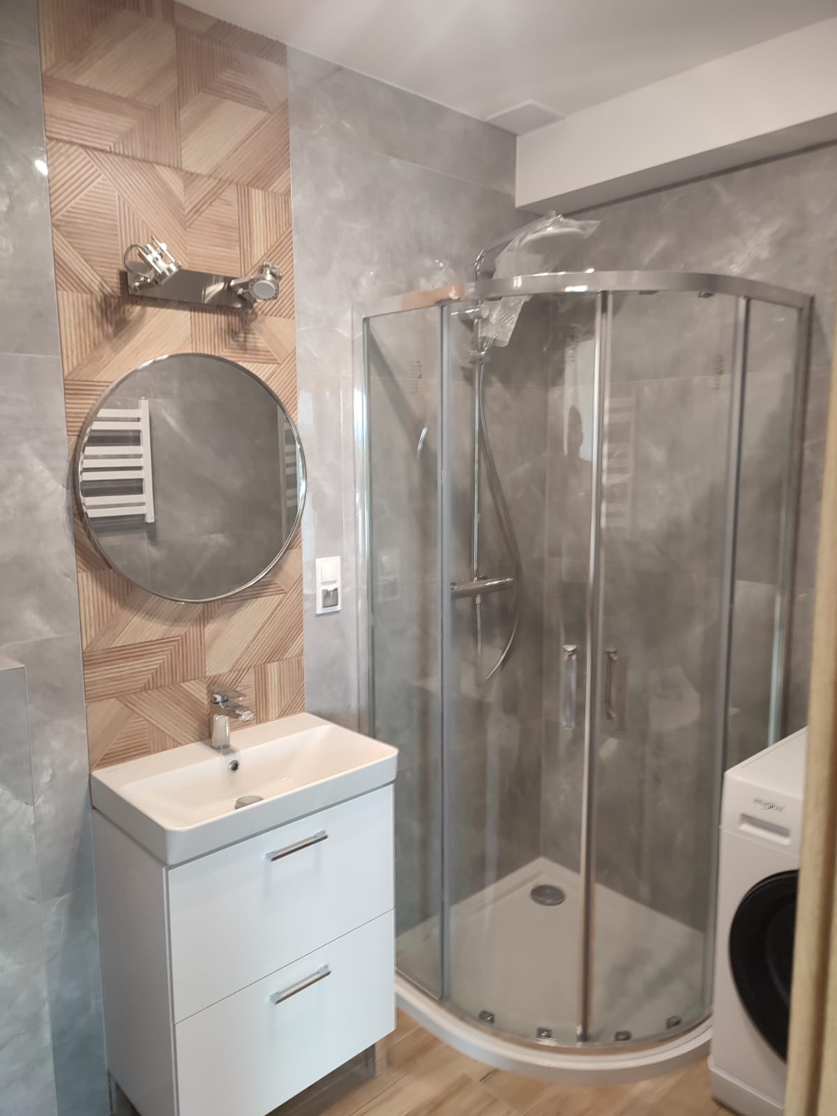 Wykończona łazienka z kabiną prysznicową narożną, umywalką z szafką w kolorze białym, okrągłym lustrem i płytkami imitującymi drewno na ścianie.