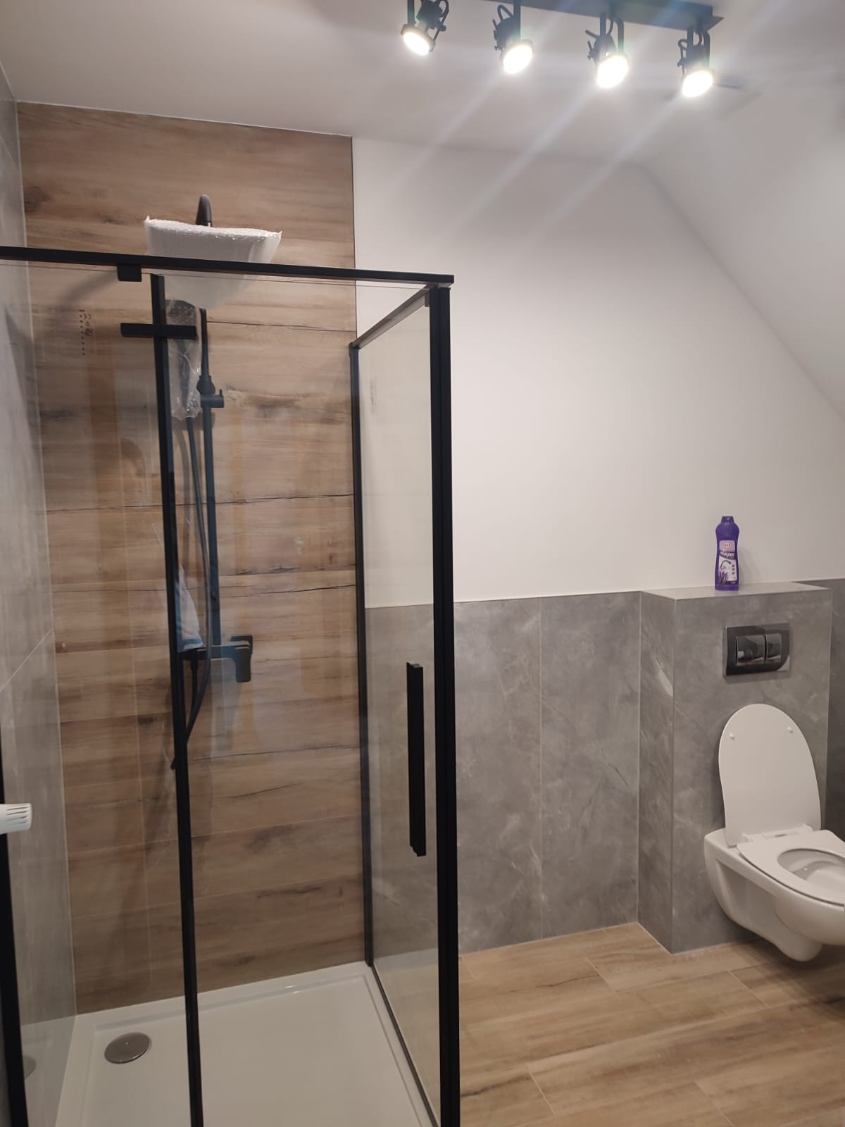 Nowoczesna łazienka z kabiną prysznicową w czarnej ramie, drewnopodobnymi płytkami na ścianie i podłodze, oraz wiszącą toaletą. Na półce nad toaletą stoi fioletowy płyn do czyszczenia.