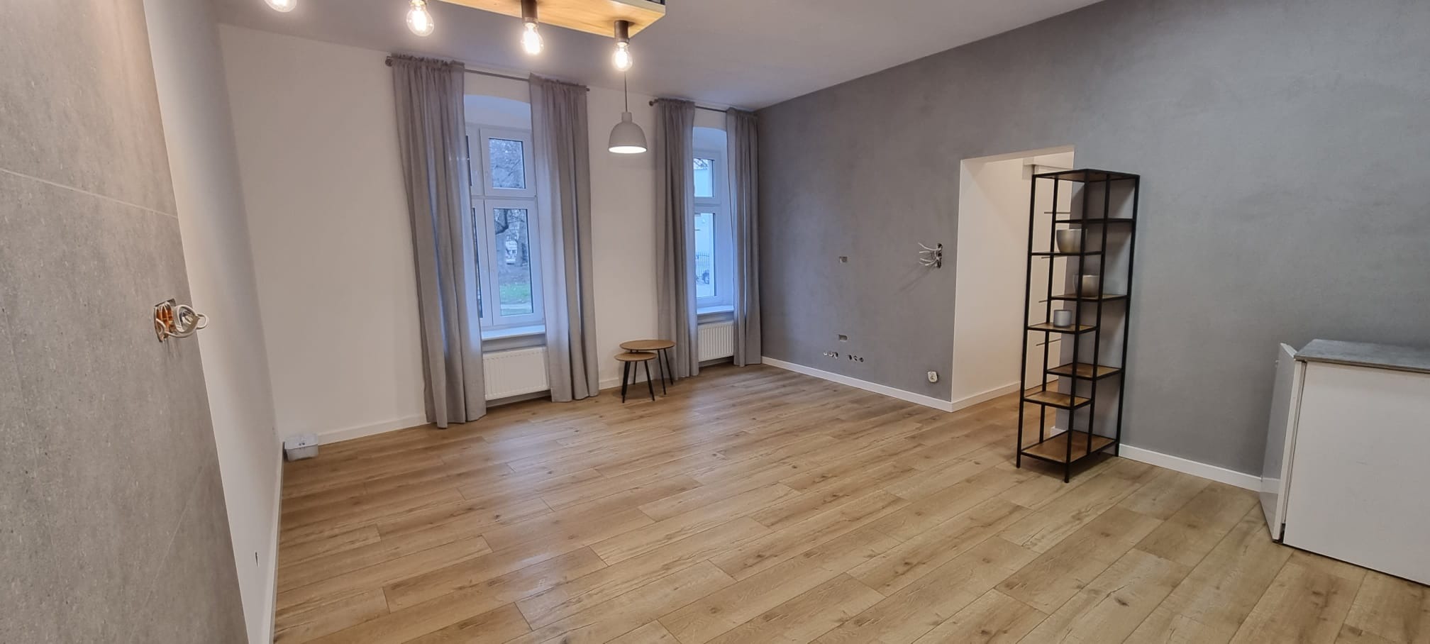 Jasne panele podłogowe w przestronnym pokoju z szarą ścianą, dwoma oknami z szarymi zasłonami, drewnianym oświetleniem sufitowym, metalowym regałem oraz małymi okrągłymi stolikami.
