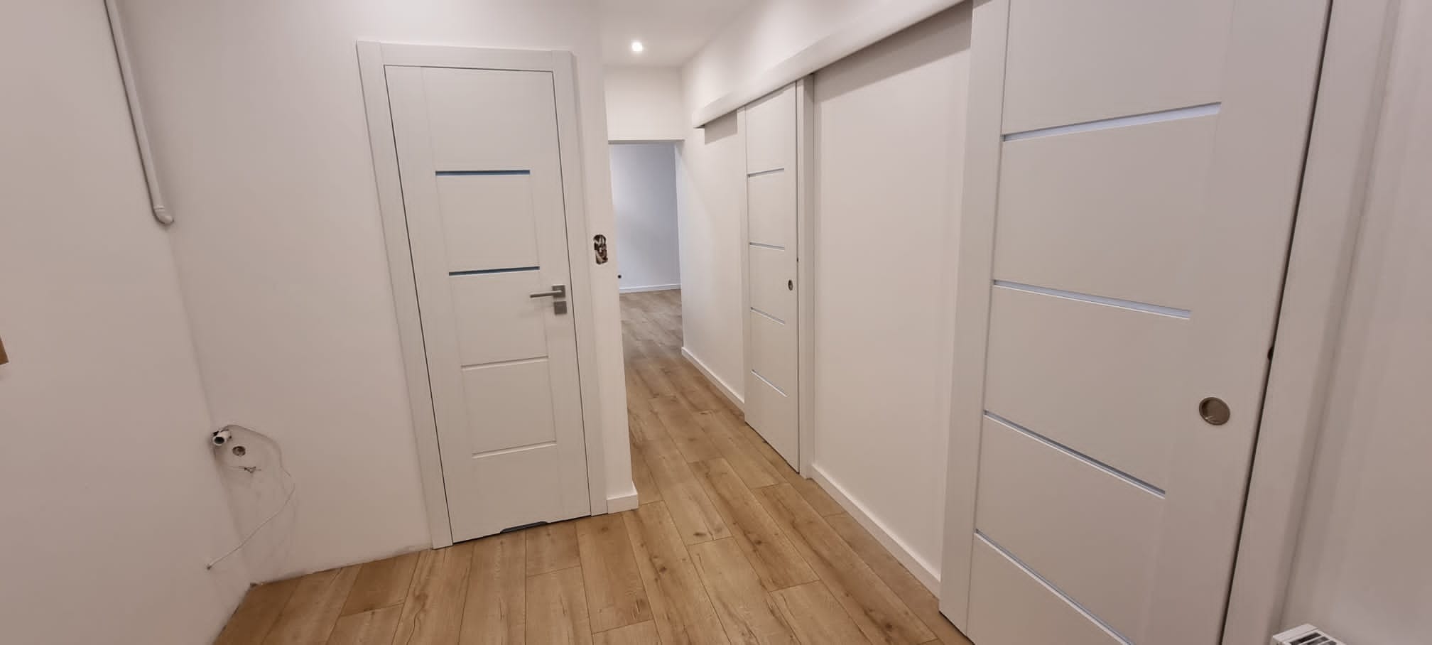 Wnętrze mieszkania po remoncie: białe ściany, panele podłogowe imitujące drewno, dwoje drzwi wewnętrznych - jedne otwarte, drugie przesuwne, widoczne gniazdka elektryczne bez osprzętu na ścianie.