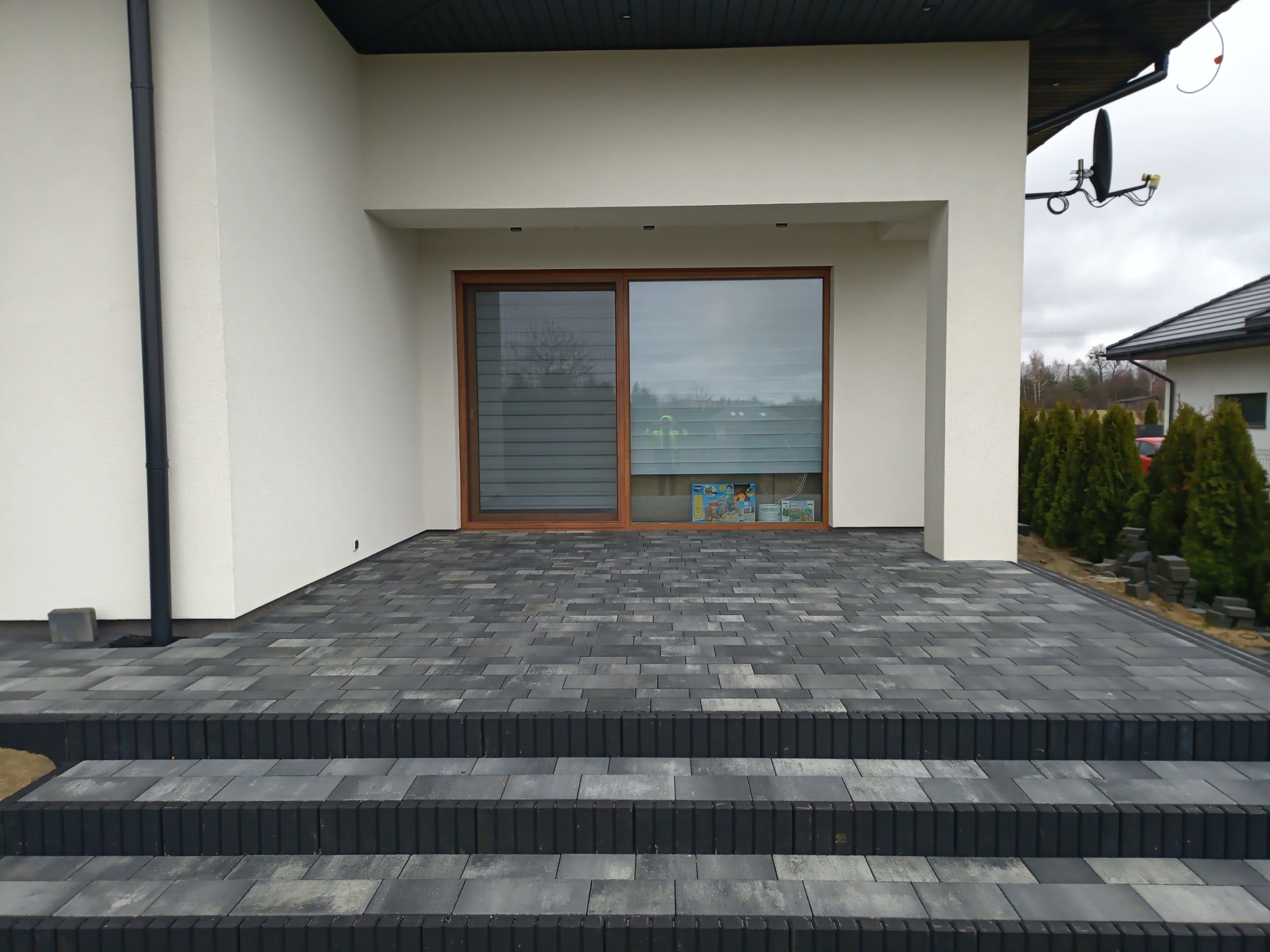 Nowoczesny taras z szarej kostki brukowej ułożonej w regularny wzór, prowadzący do dużego okna domu z widocznymi roletami i pudełkami z zabawkami wewnątrz.