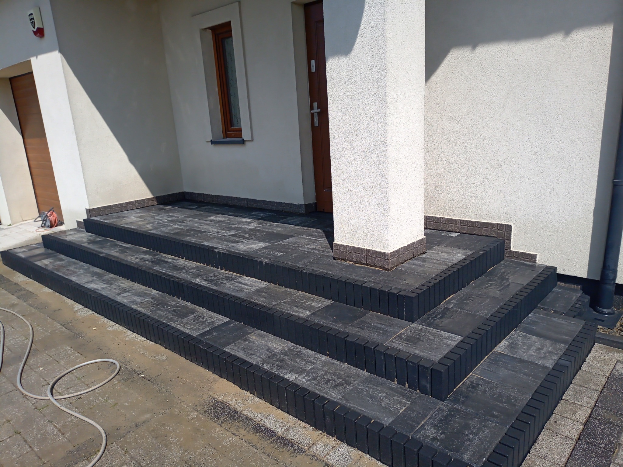 Schody wejściowe do domu wyłożone ciemną kostką brukową z obrzeżami, widoczne drzwi wejściowe i fragment elewacji budynku.