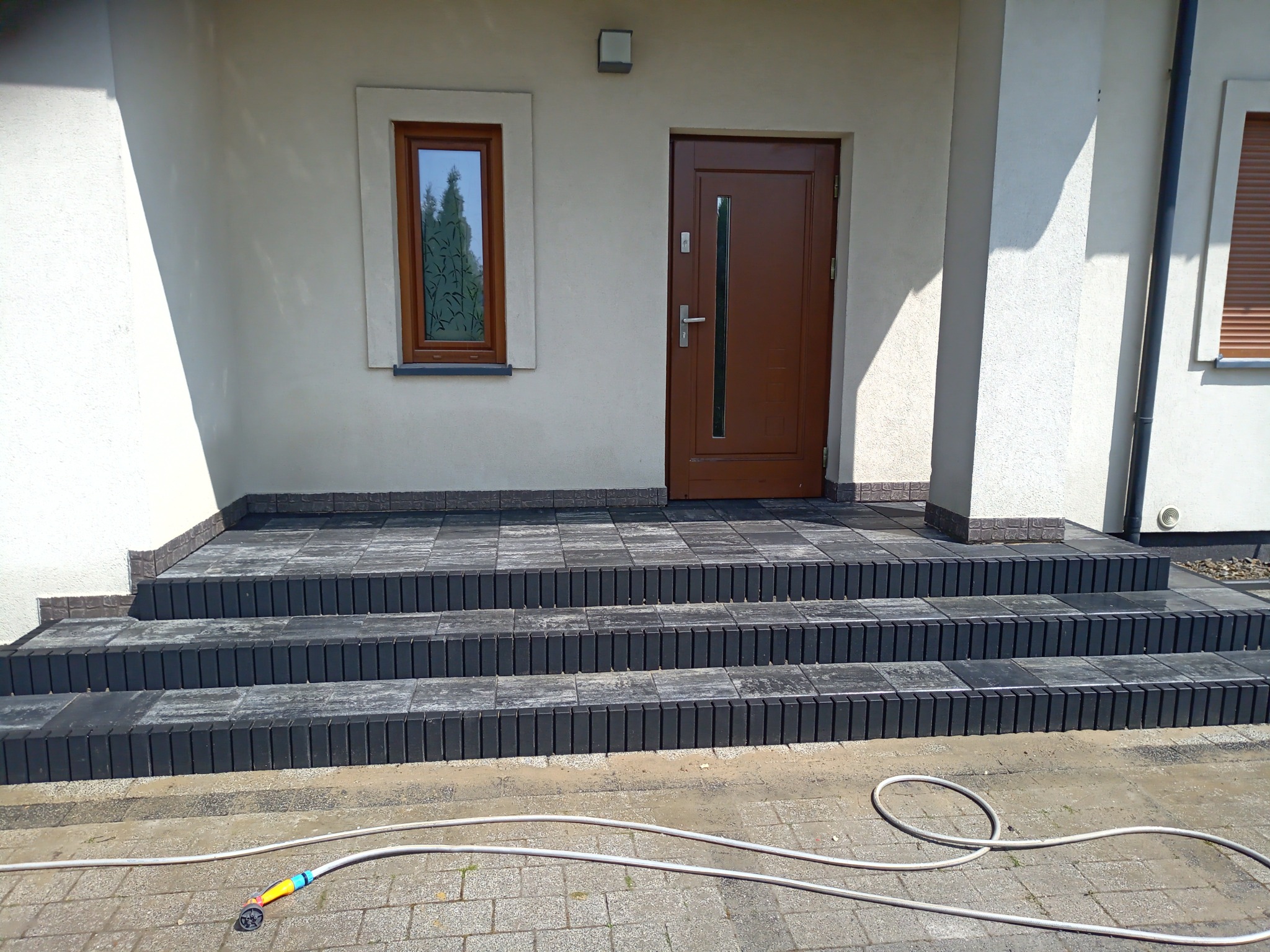 Schody wejściowe do domu wyłożone szarą kostką brukową, widoczny wąż ogrodowy z dyszą na kostce przed schodami.