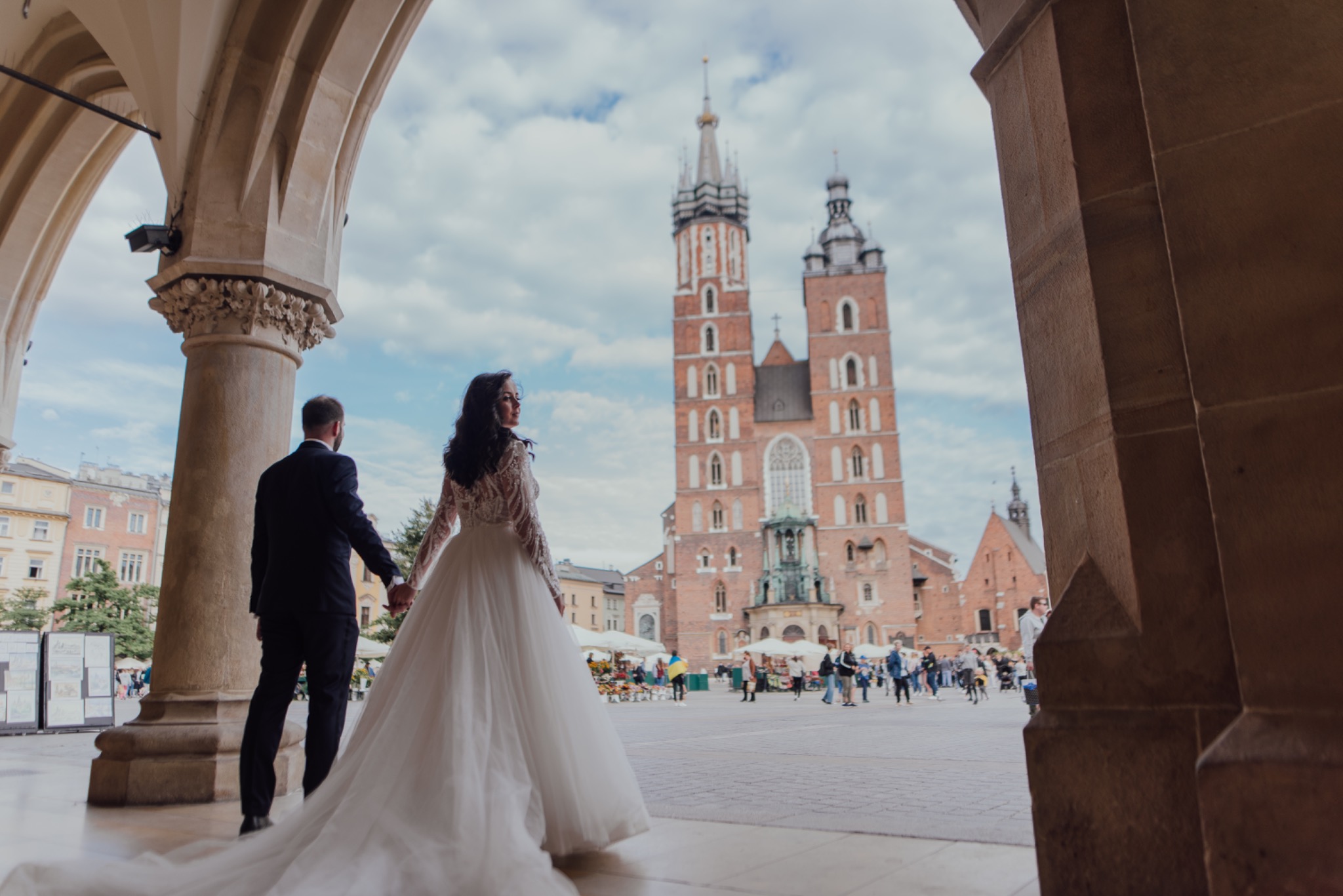 Para młoda trzymająca się za ręce, spacerująca pod arkadami Sukiennic w Krakowie, z widokiem na Bazylikę Mariacką w tle.
