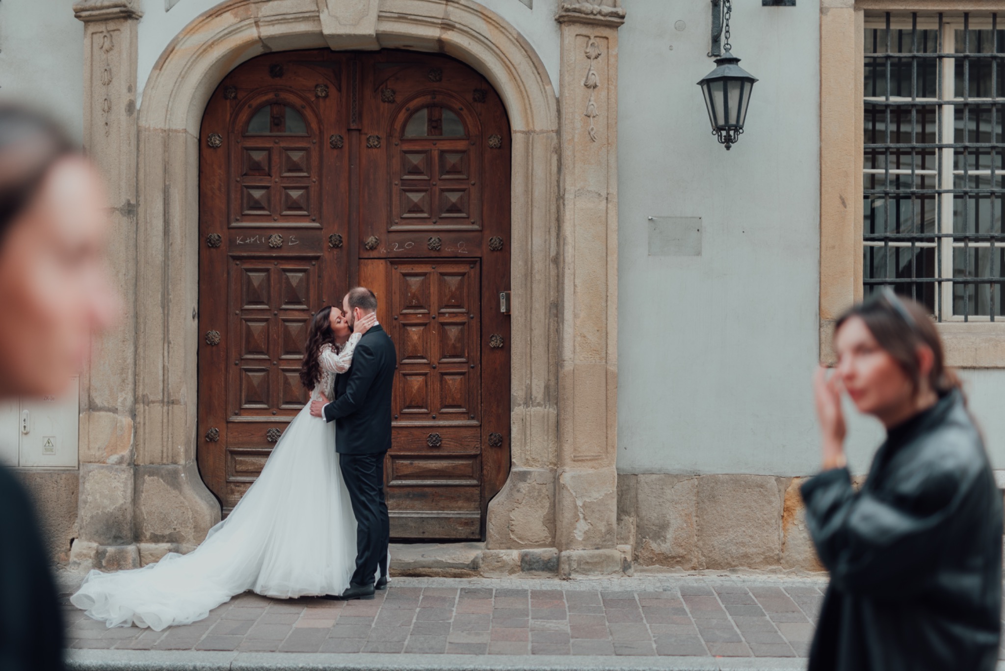 Pocałunek pary młodej na tle zabytkowych drewnianych drzwi z metalowymi okuciami, w tle fragmenty twarzy dwóch osób.