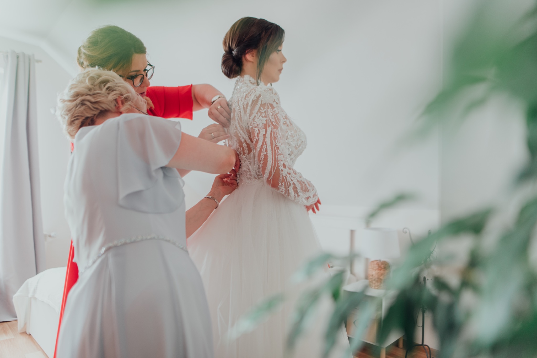 Panna młoda w koronkowej sukni ślubnej z długim rękawem, zapinana przez dwie kobiety w jasnych sukienkach w jasnym pokoju.