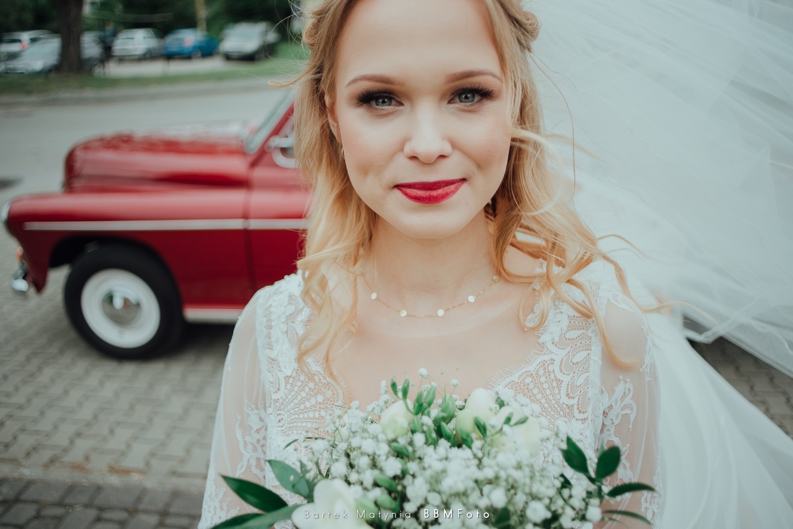 Portret uśmiechniętej panny młodej z bukietem na tle czerwonego, klasycznego samochodu i rozwianego welonu.