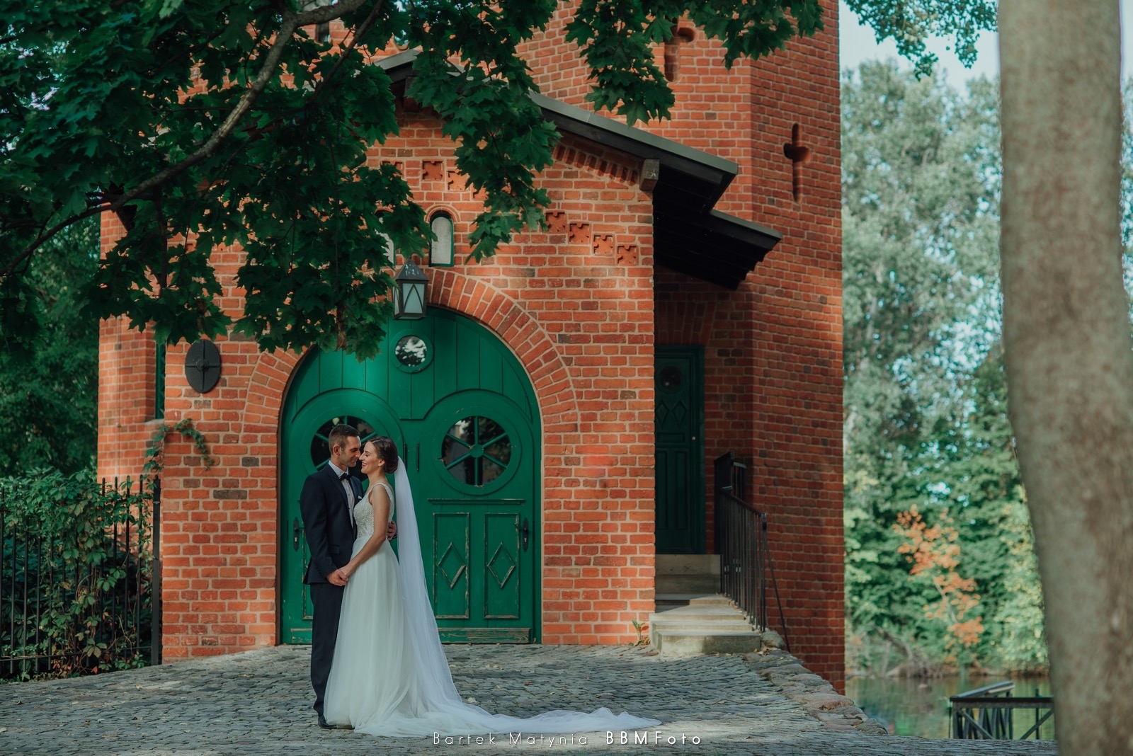Para młoda w objęciach na tle ceglanej budowli z zielonymi drzwiami łukowymi i brukowanym chodnikiem, w tle drzewa.