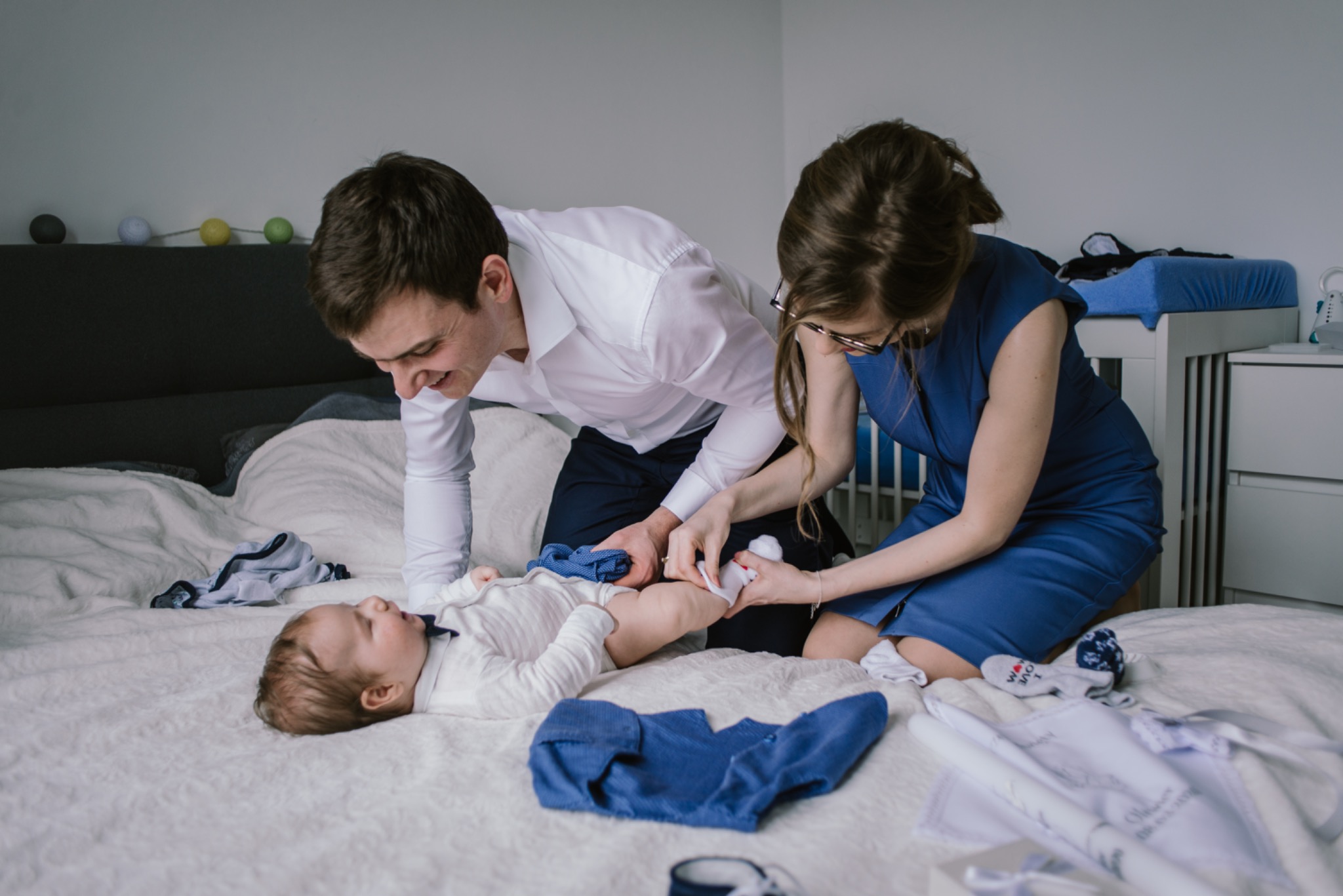 Rodzice przebierają niemowlę leżące na łóżku, w tle dziecięce łóżeczko i girlanda z kolorowych kul.