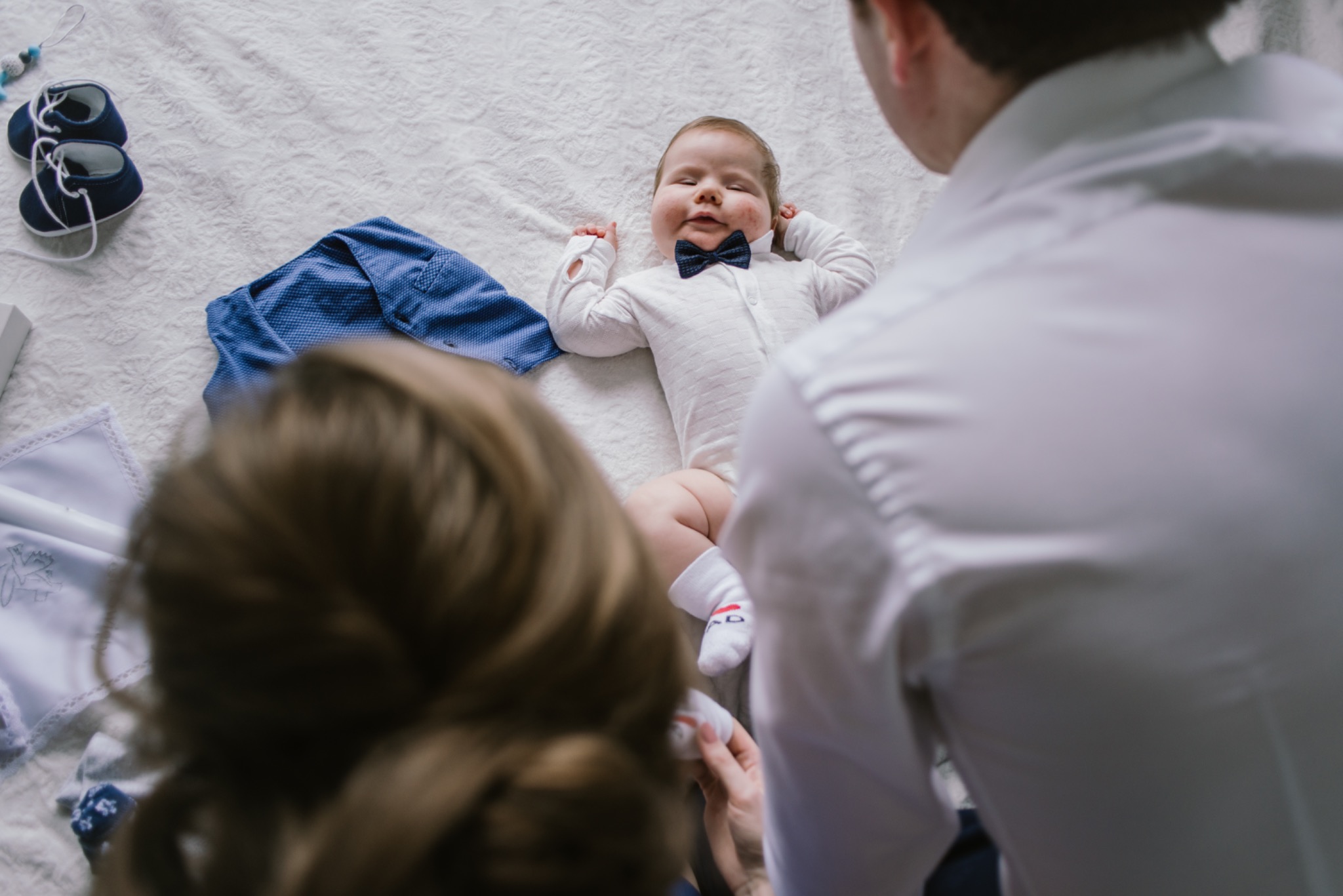 Uśmiechnięty niemowlak w białym body i granatowej muszce leży na białym kocu, obok leżą ubranka i buciki, rodzice w tle.