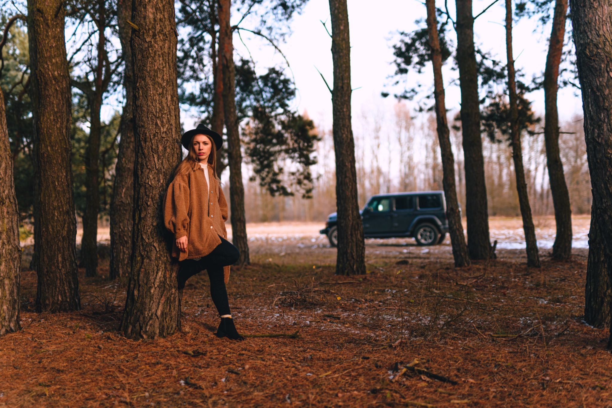 Kobieta w brązowej kurtce i kapeluszu pozuje oparta o drzewo w lesie, w tle widoczny samochód terenowy.