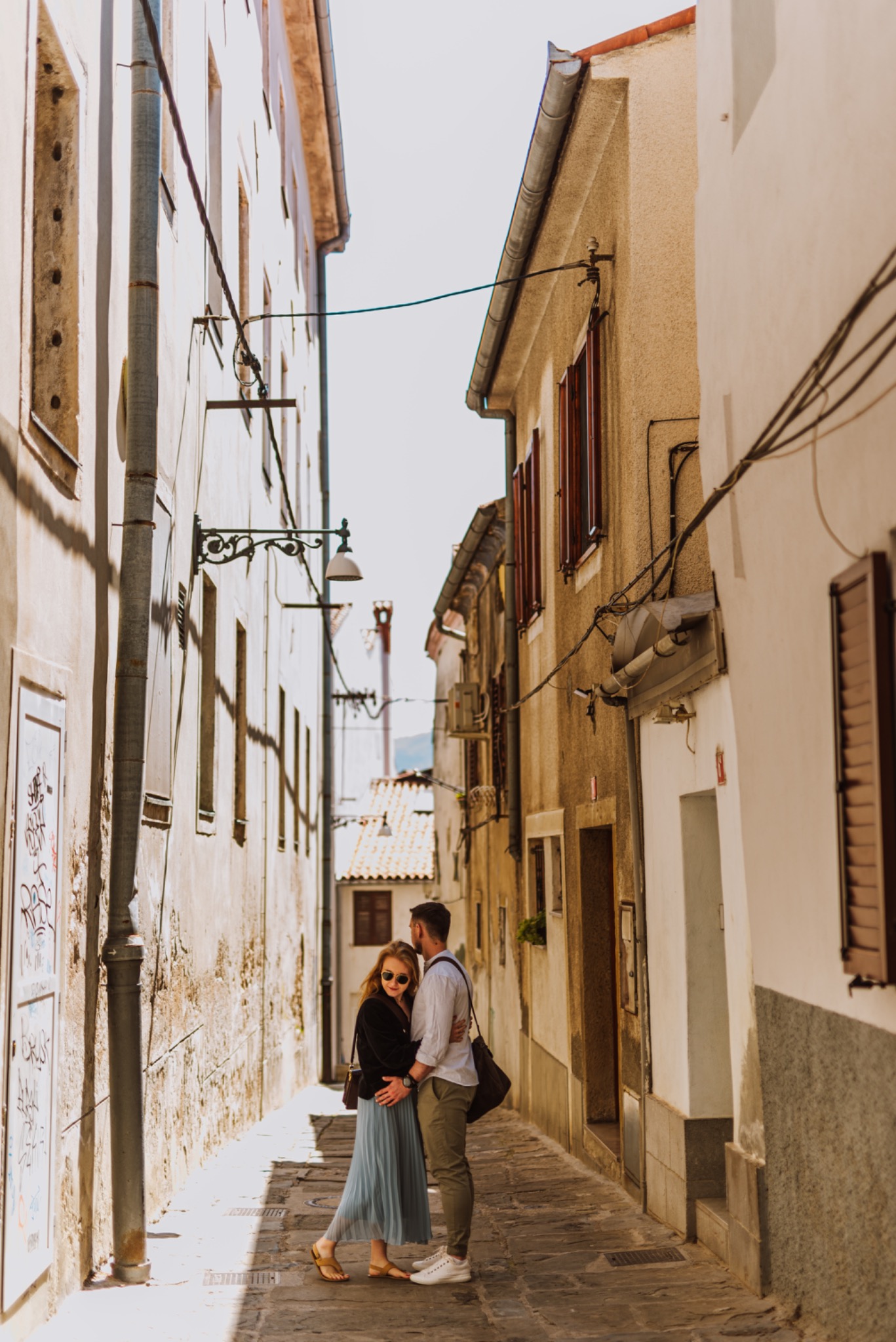 Para zakochanych w objęciach na wąskiej, słonecznej uliczce z kamienną nawierzchnią i budynkami o zróżnicowanej architekturze, kobieta w zwiewnej, błękitnej spódnicy i ciemnej kurtce, mężczyzna...