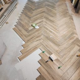 WYKOŃCZENIE WNĘTRZ DENYS POLTAVETS - Układanie drewnopodobnych płytek w jodełkę na podłodze, widoczne białe krzyżyki dystansowe, zielone klipsy poziomujące i pędzel do rozprowadzania fugi.