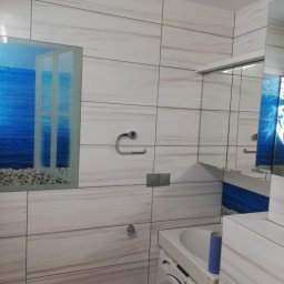 WYKOŃCZENIE WNĘTRZ DENYS POLTAVETS - Biała łazienka w Skowarczu z płytkami imitującymi marmur, lustro z motywem morskim i dekoracyjnym oknem z widokiem na morze, oraz widoczna pralka.