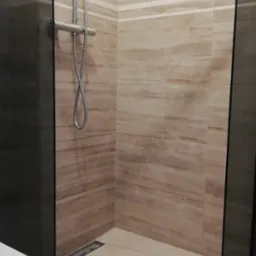 Kabina prysznicowa walk-in z odpływem liniowym, wykończona płytkami imitującymi marmur w odcieniach beżu i brązu, widoczny fragment toalety.