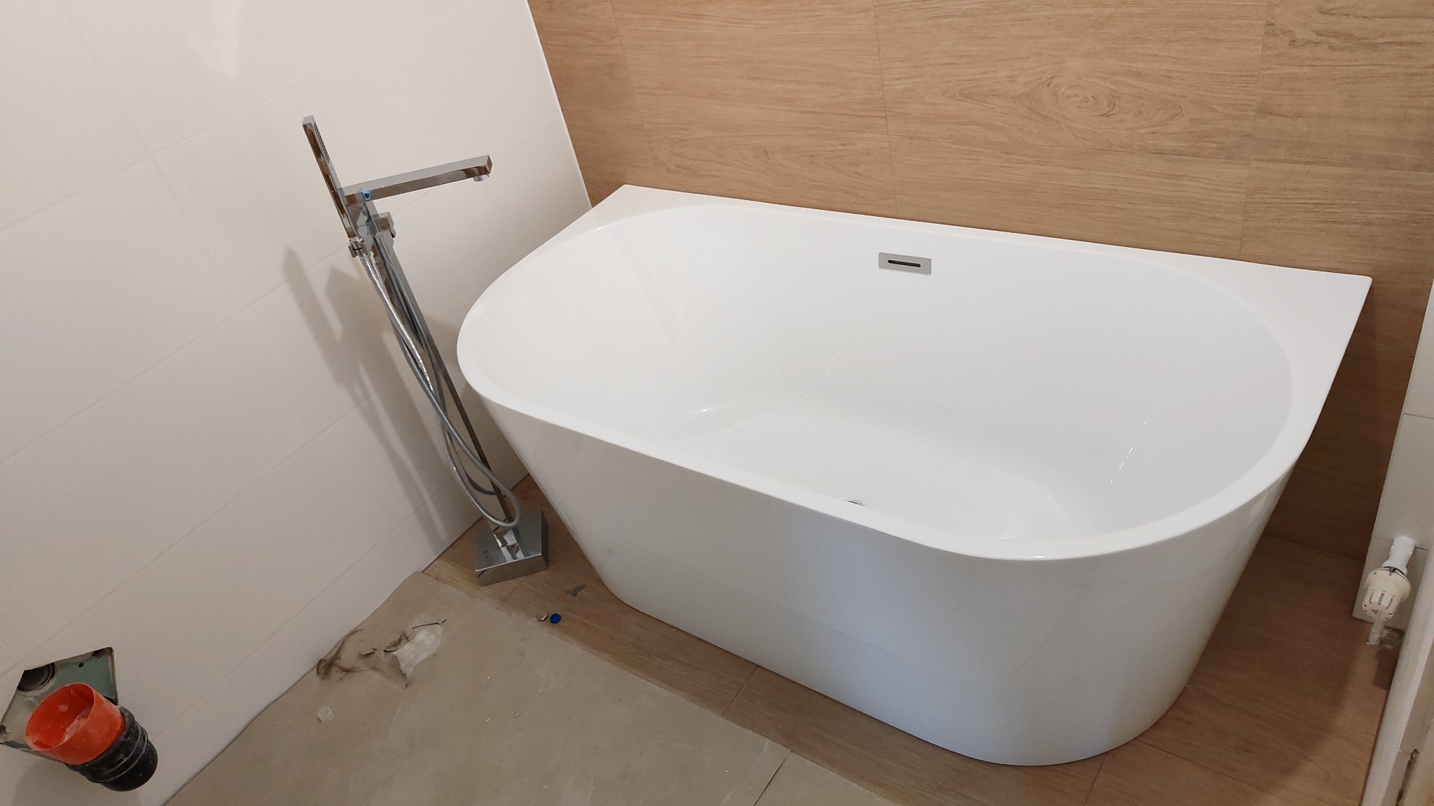 Biała, asymetryczna wanna wolnostojąca z chromowaną baterią podłogową, zamontowana przy ścianie wyłożonej białymi płytkami; w tle widoczna ściana z płytkami imitującymi drewno, na podłodze widoczne...