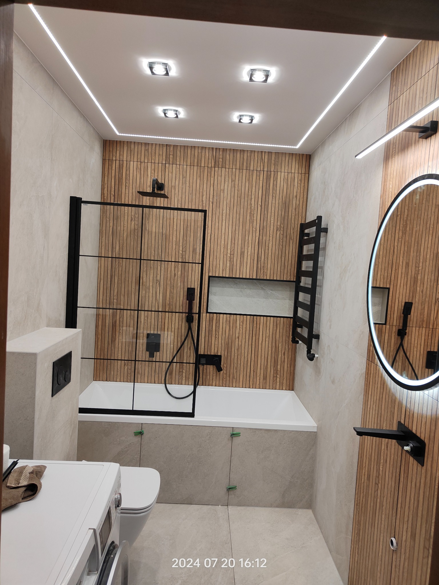 Nowoczesna łazienka z drewnianymi panelami ściennymi, czarną armaturą i oświetleniem LED, widok na wannę za czarną szybą prysznicową, okrągłe lustro z podświetleniem i pralkę z ręcznikiem.