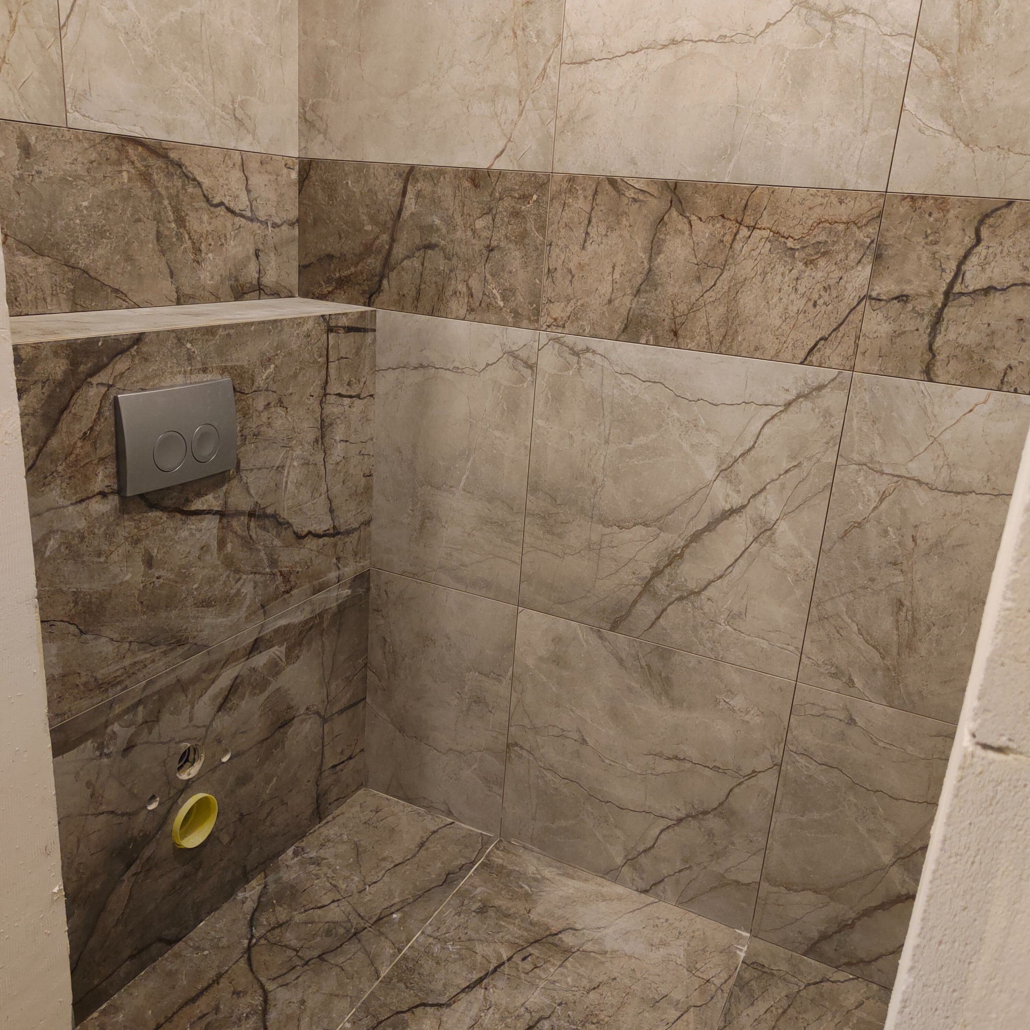 Fragment łazienki z marmurowymi płytkami w odcieniach beżu i brązu, widoczny stelaż podtynkowy WC z szarym panelem przycisków, otwory instalacyjne w ścianie.