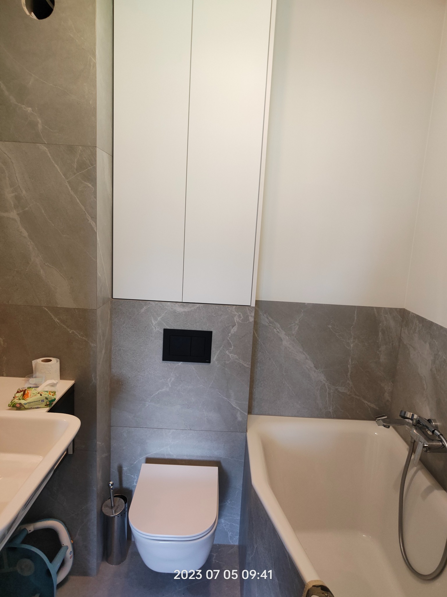 Nowoczesna łazienka z szarymi płytkami imitującymi marmur, białą zabudową nad toaletą i wanną, oraz widoczną umywalką. Na blacie przy umywalce papier toaletowy i chusteczki. Widoczny fragment...