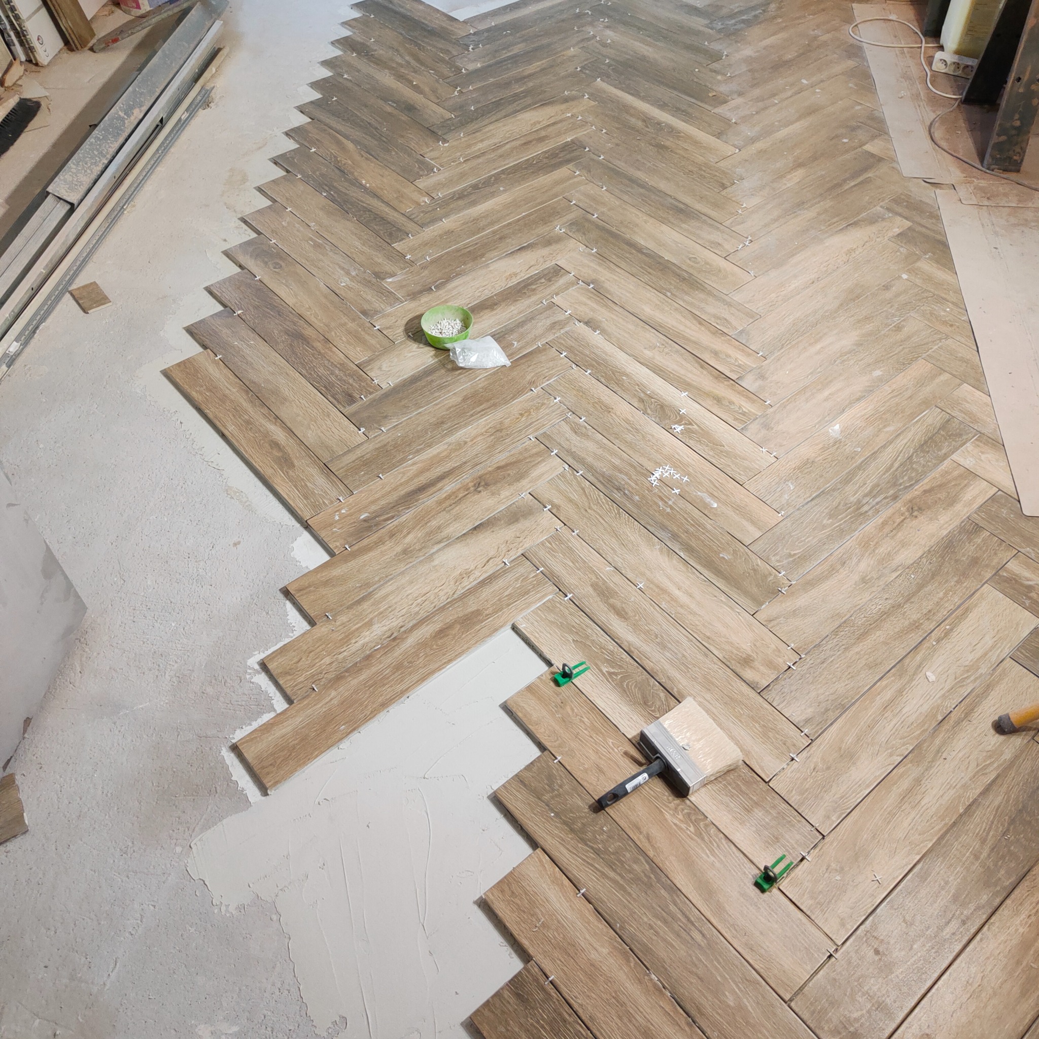 Układanie drewnopodobnych płytek w jodełkę na podłodze, widoczne białe krzyżyki dystansowe, zielone klipsy poziomujące i pędzel do rozprowadzania fugi.