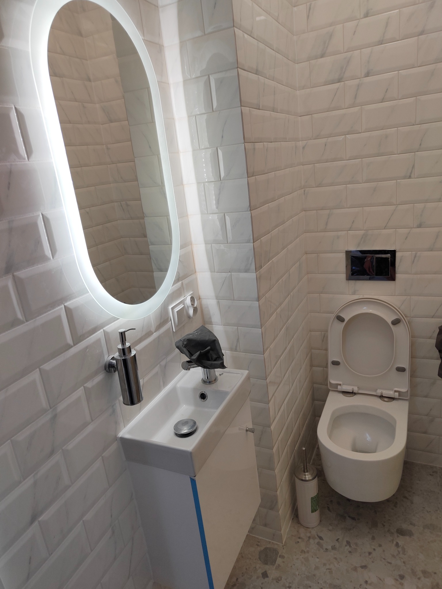 Nowoczesna, mała łazienka z białymi płytkami imitującymi cegłę, podświetlanym lustrem o owalnym kształcie, wiszącą umywalką z szafką, toaletą i podłogą z lastryko.