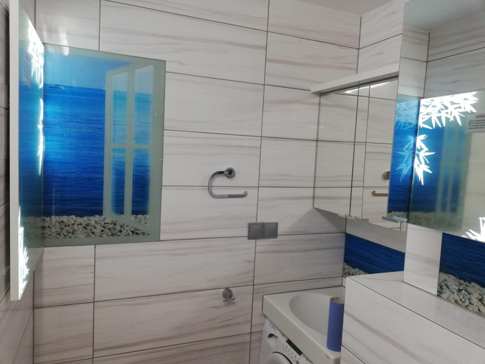 Biała łazienka w Skowarczu z płytkami imitującymi marmur, lustro z motywem morskim i dekoracyjnym oknem z widokiem na morze, oraz widoczna pralka.