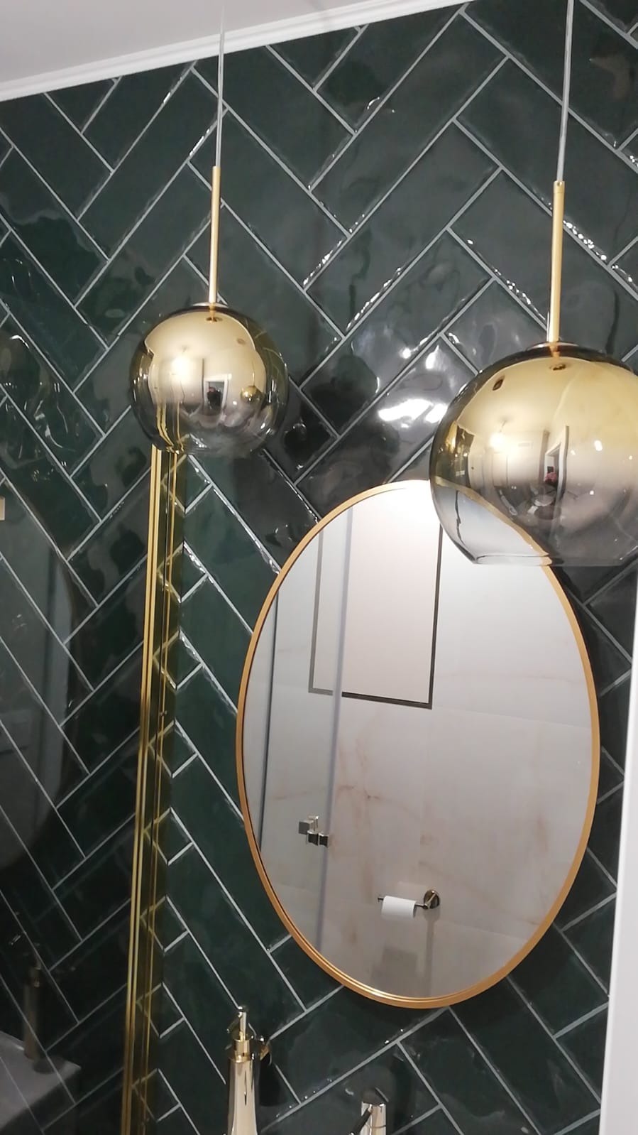 Luksusowa łazienka z ciemnozielonymi płytkami ułożonymi w jodełkę, okrągłym lustrem w złotej ramie i wiszącymi lampami w kształcie złotych kul.