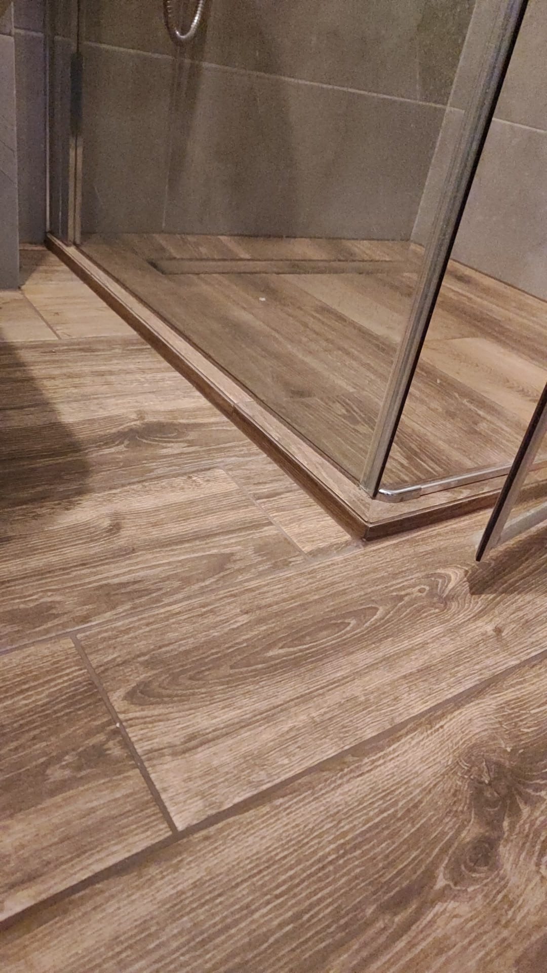 Szczegółowe ujęcie podłogi w łazience wyłożonej płytkami imitującymi drewno, z widocznym fragmentem kabiny prysznicowej z metalowym obramowaniem.
