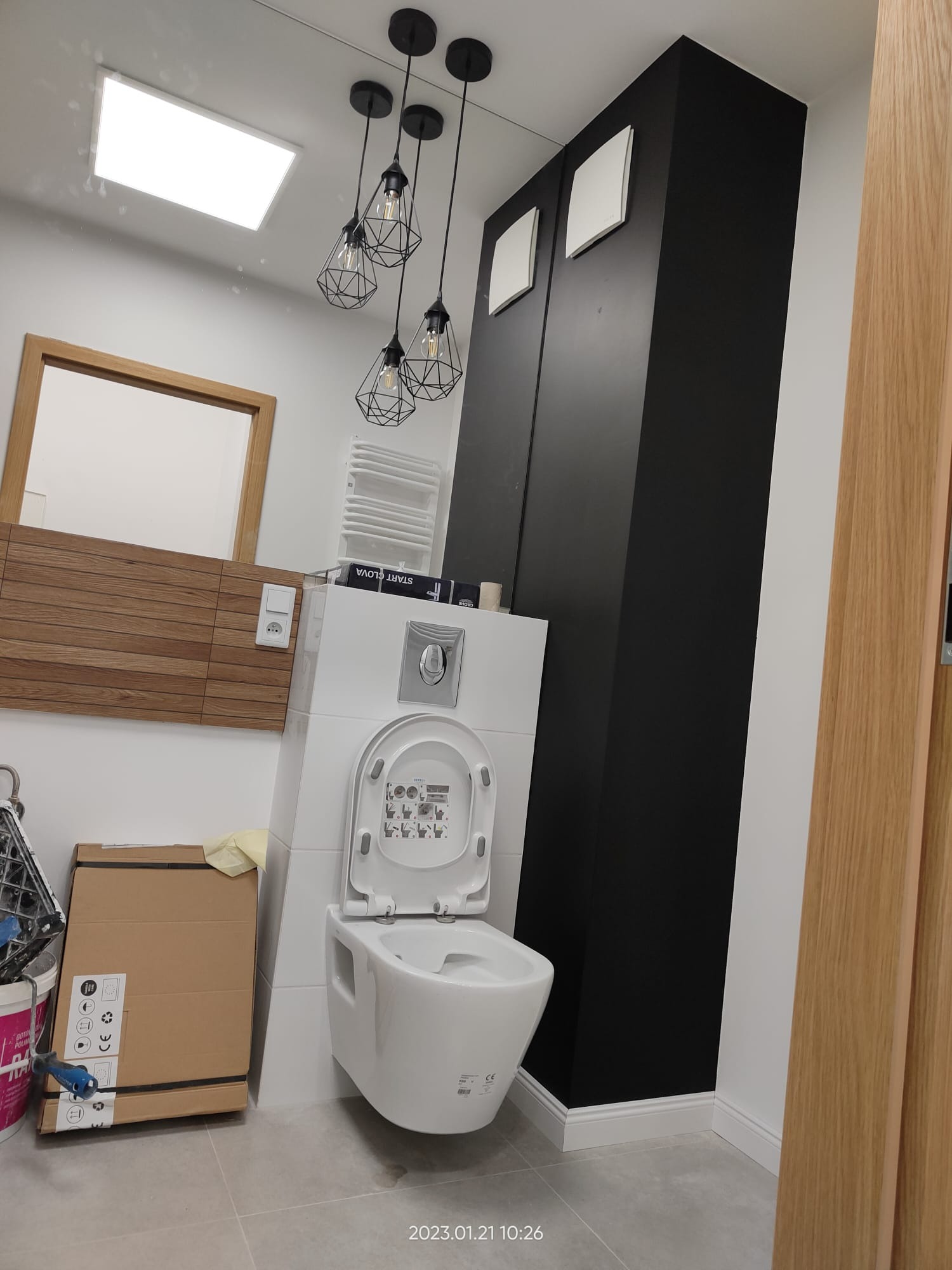 Nowoczesna łazienka z podwieszaną toaletą z otwartą klapą, czarnym wysokim regałem z wentylacją, drewnianą boazerią i designerskimi lampami wiszącymi na tle białych płytek.