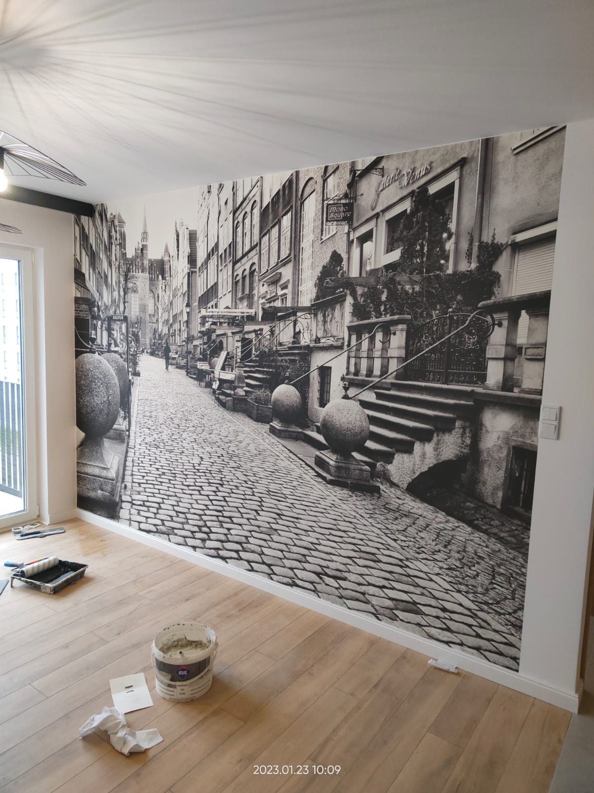 Czarno-biała fototapeta przedstawiająca uliczkę starego miasta, z widocznym wiadrem z klejem i wałkiem malarskim na podłodze z paneli.