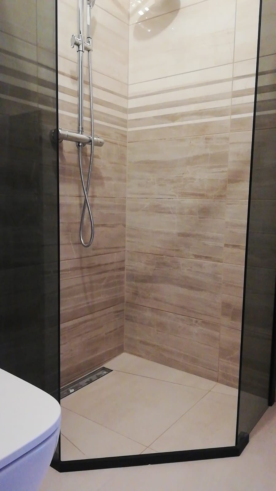 Kabina prysznicowa walk-in z odpływem liniowym, wykończona płytkami imitującymi marmur w odcieniach beżu i brązu, widoczny fragment toalety.