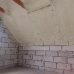 Poddasze w trakcie ocieplania: ściany z bloczków betonowych, widoczne belki stropowe i nałożona warstwa izolacji natryskowej z pianki.